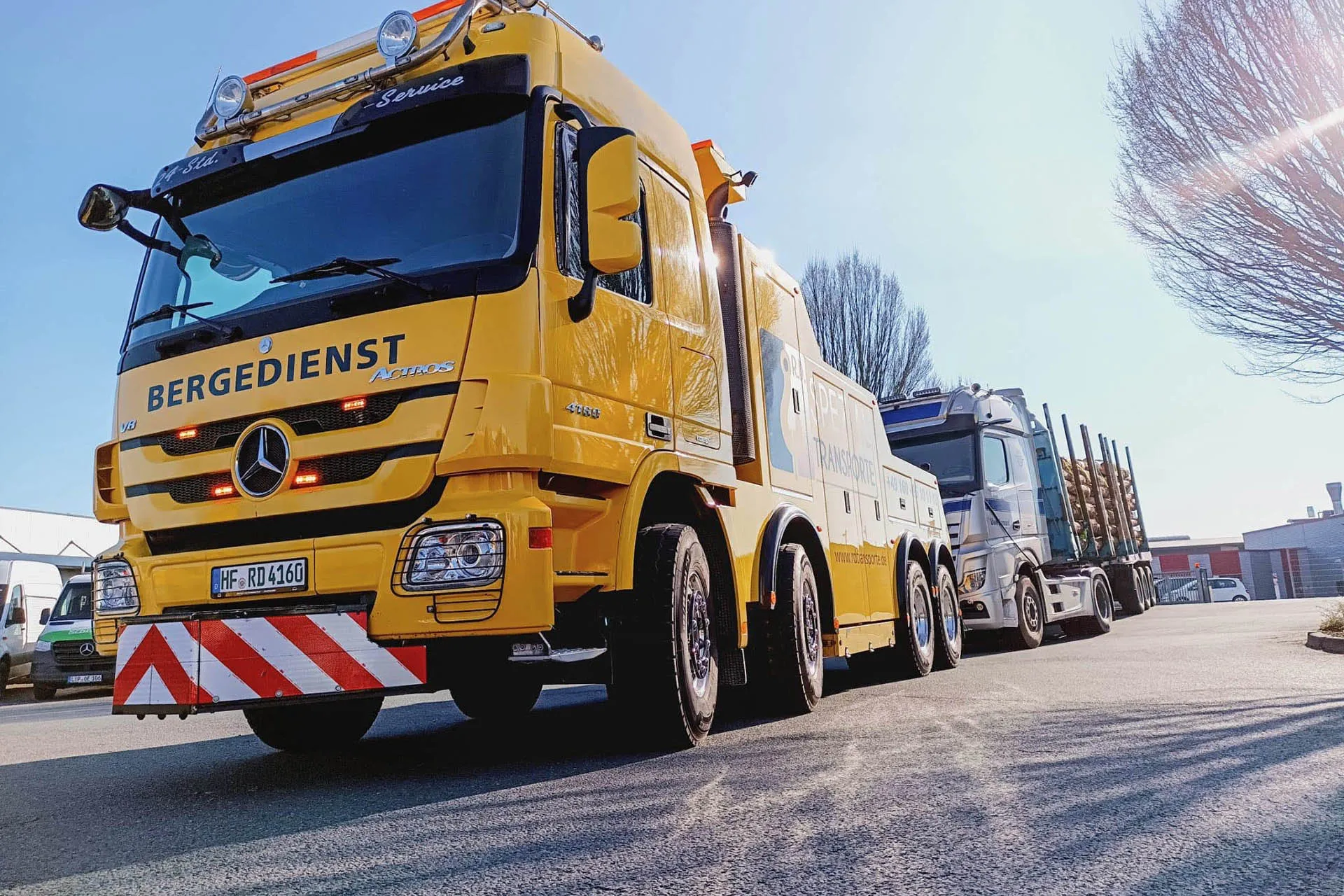 Gelber Mercedes-Benz Actros Bergedienst-LKW von R.D. Spezial-Transporte im Einsatz, frontal von unten fotografiert, mit einem Holztransporter im Hintergrund bei Sonnenlicht.