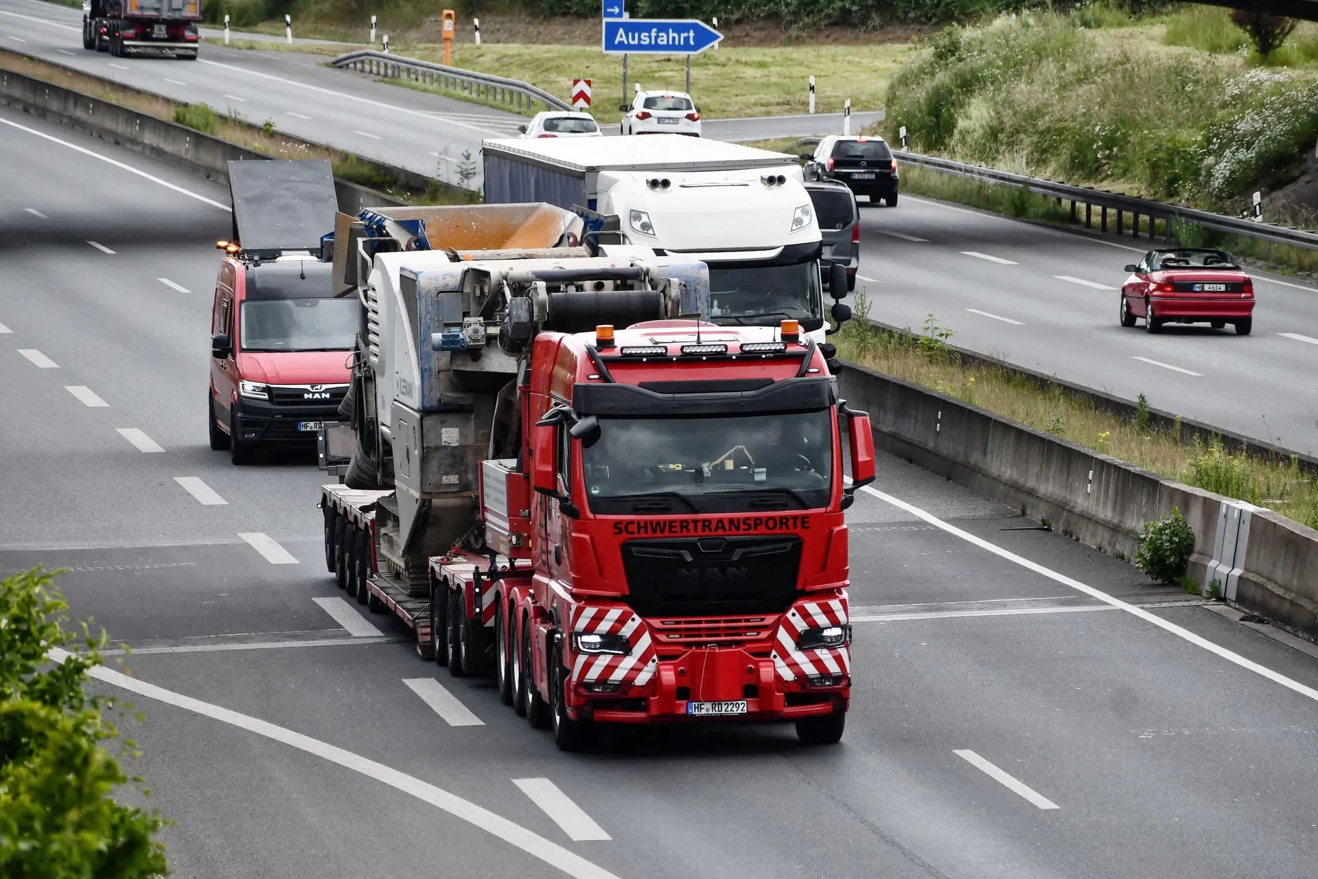 Roter MAN Schwertransporter von R.D. Spezial-Transporte mit einer großen Industriemaschine auf der Ladefläche fährt auf einer mehrspurigen Autobahn, begleitet von einem Servicefahrzeug.