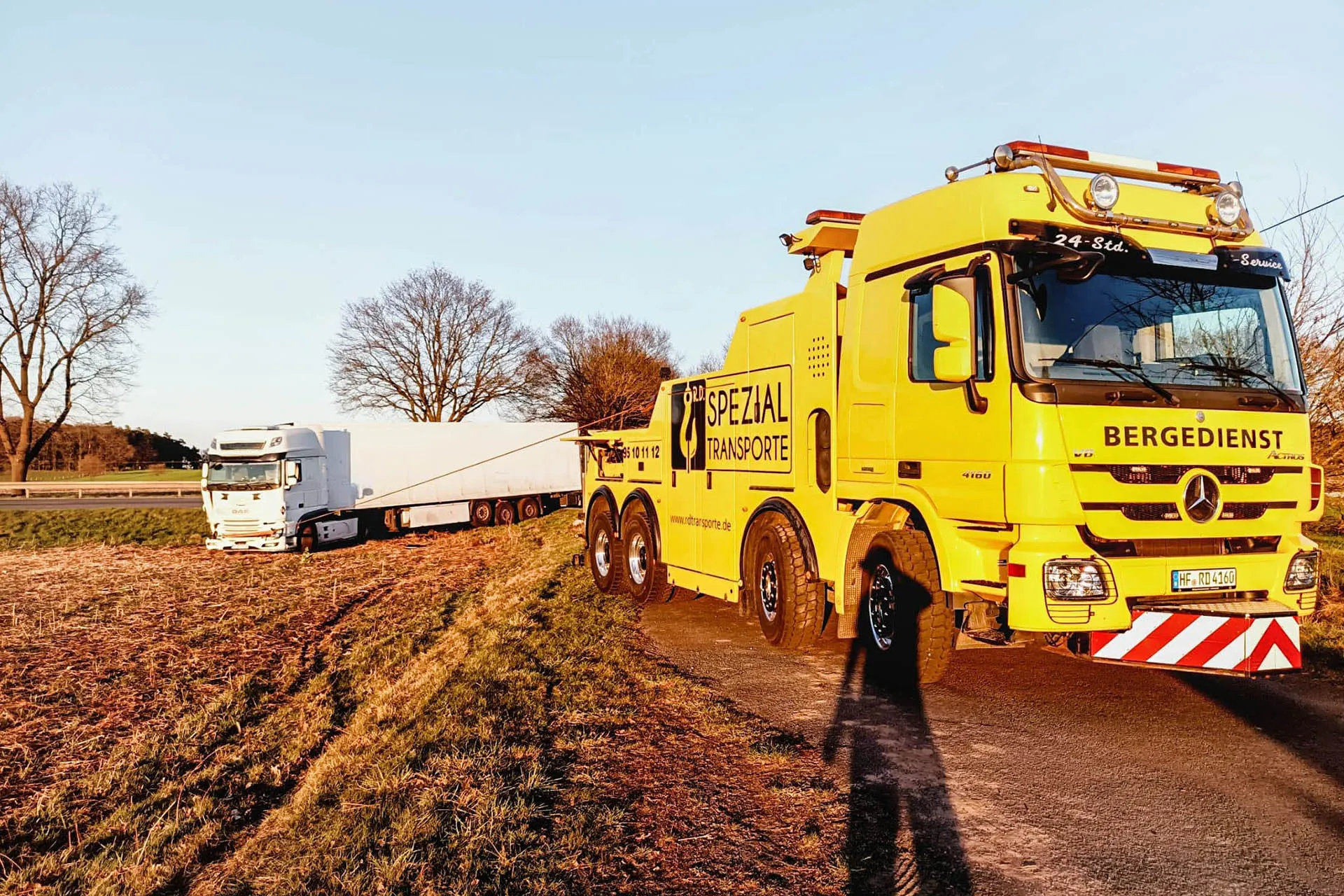 Gelber Mercedes-Benz Bergedienst-LKW von R.D. Spezial-Transporte zieht einen weißen Sattelzug aus einem aufgeweichten Acker am Straßenrand.