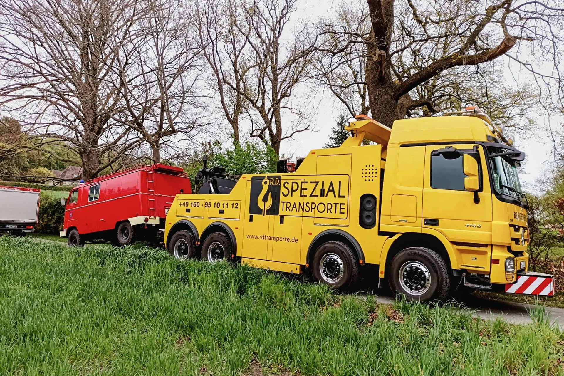 Gelber Spezial-Abschleppwagen von R.D. Spezial-Transporte zieht einen roten historischen Feuerwehrwagen auf einer von Bäumen gesäumten Straße am Wiesenrand.
