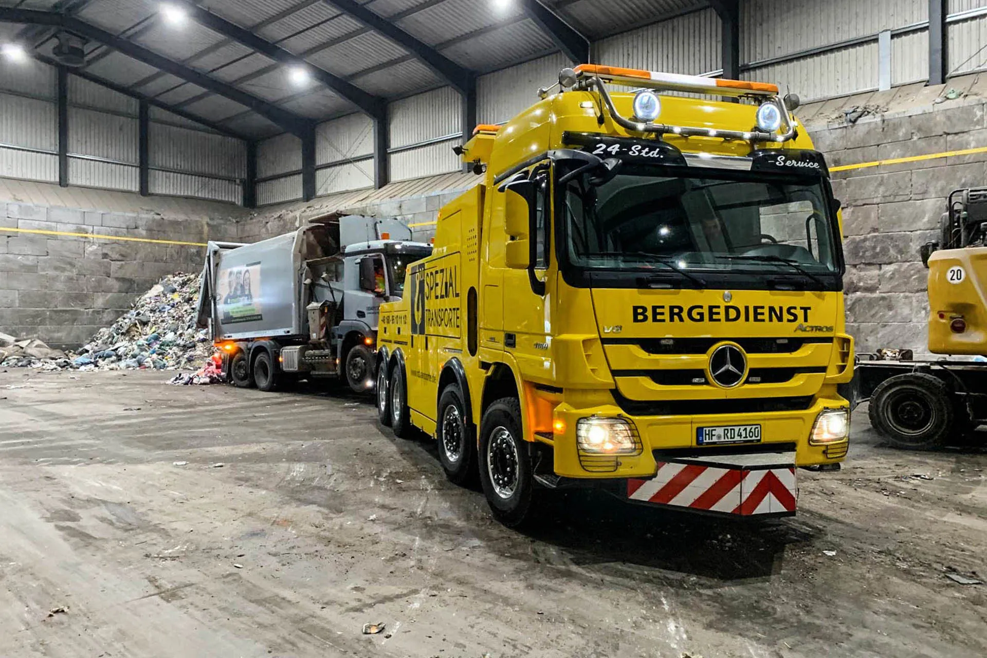 Gelber Mercedes-Benz Actros Bergedienst-LKW von R.D. Spezial-Transporte zieht einen defekten Müllwagen aus einer großen Halle mit Müllbergen.