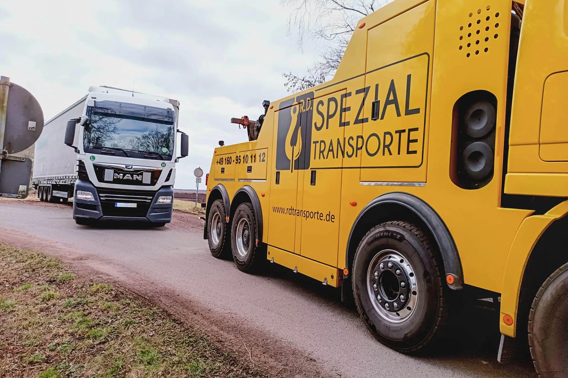 Gelber Spezial-Abschleppwagen von R.D. Spezial-Transporte fährt auf einer Landstraße hinter einem weißen MAN Sattelzug.