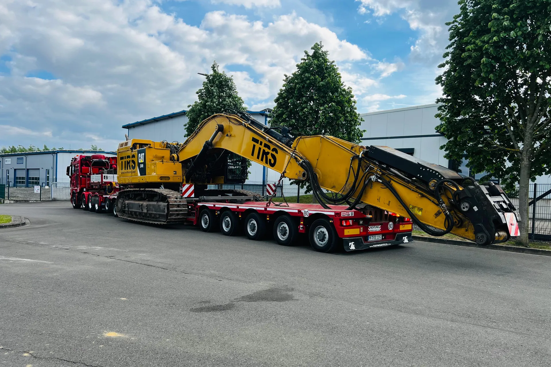 Seitliche Heckansicht eines Schwertransports von R.D. Spezial-Transporte, der einen großen gelben CAT 374F Abrissbagger auf einem roten Tieflader transportiert.