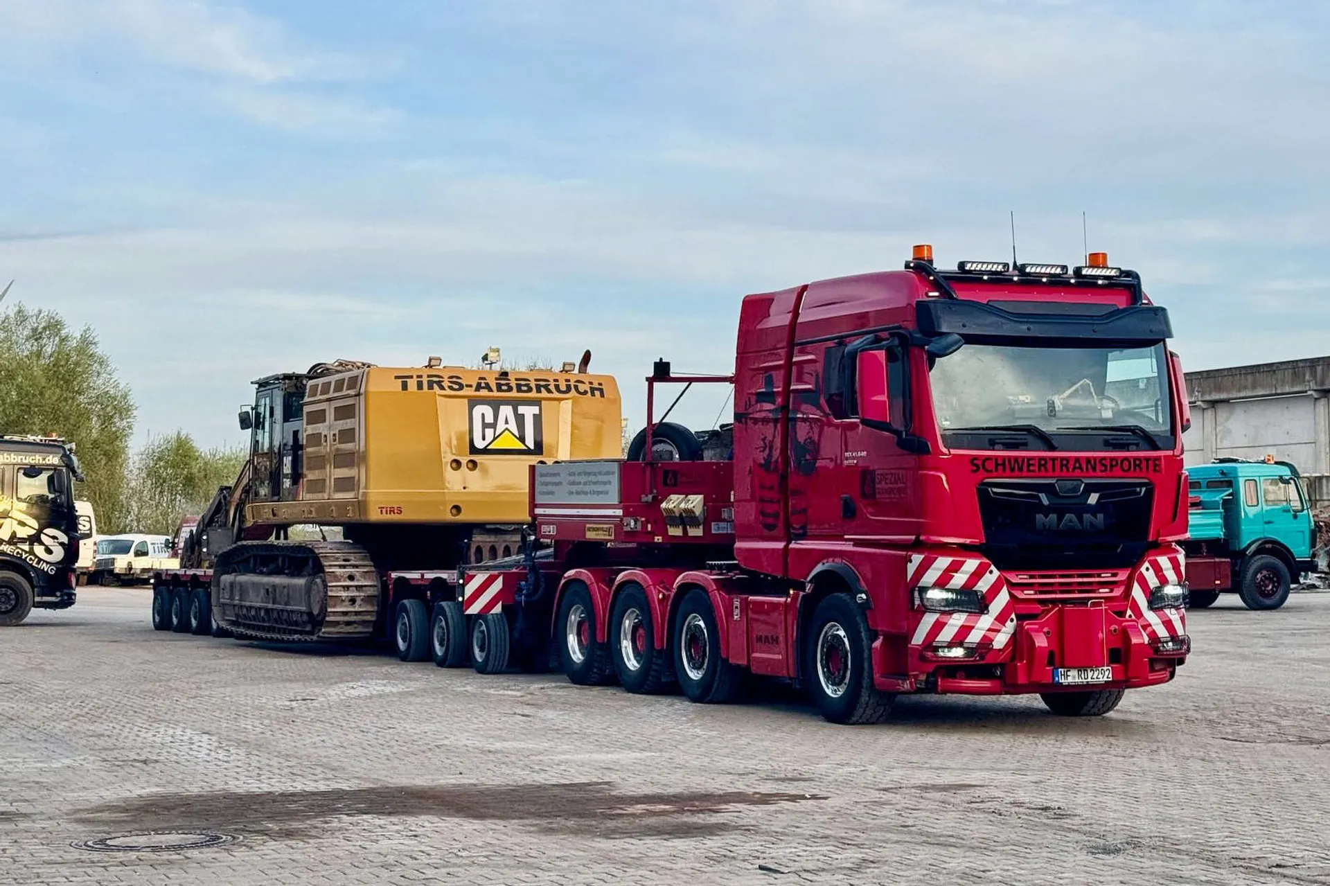 Roter MAN Schwertransporter von R.D. Spezial-Transporte transportiert einen großen gelben CAT Abrissbagger auf einem mehrachsigen Tieflader auf einem Betriebsgelände.