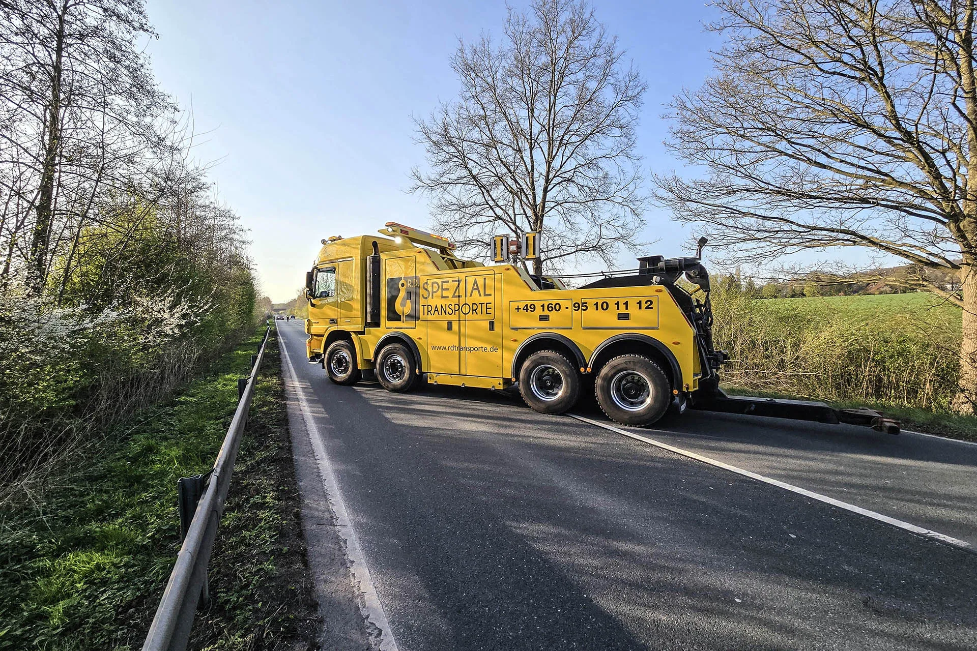 Gelber Scania Spezial-Abschleppwagen von R.D. Spezial-Transporte mit ausgefahrener Abschleppbrille auf einer Landstraße, gesäumt von Bäumen und blühenden Büschen.