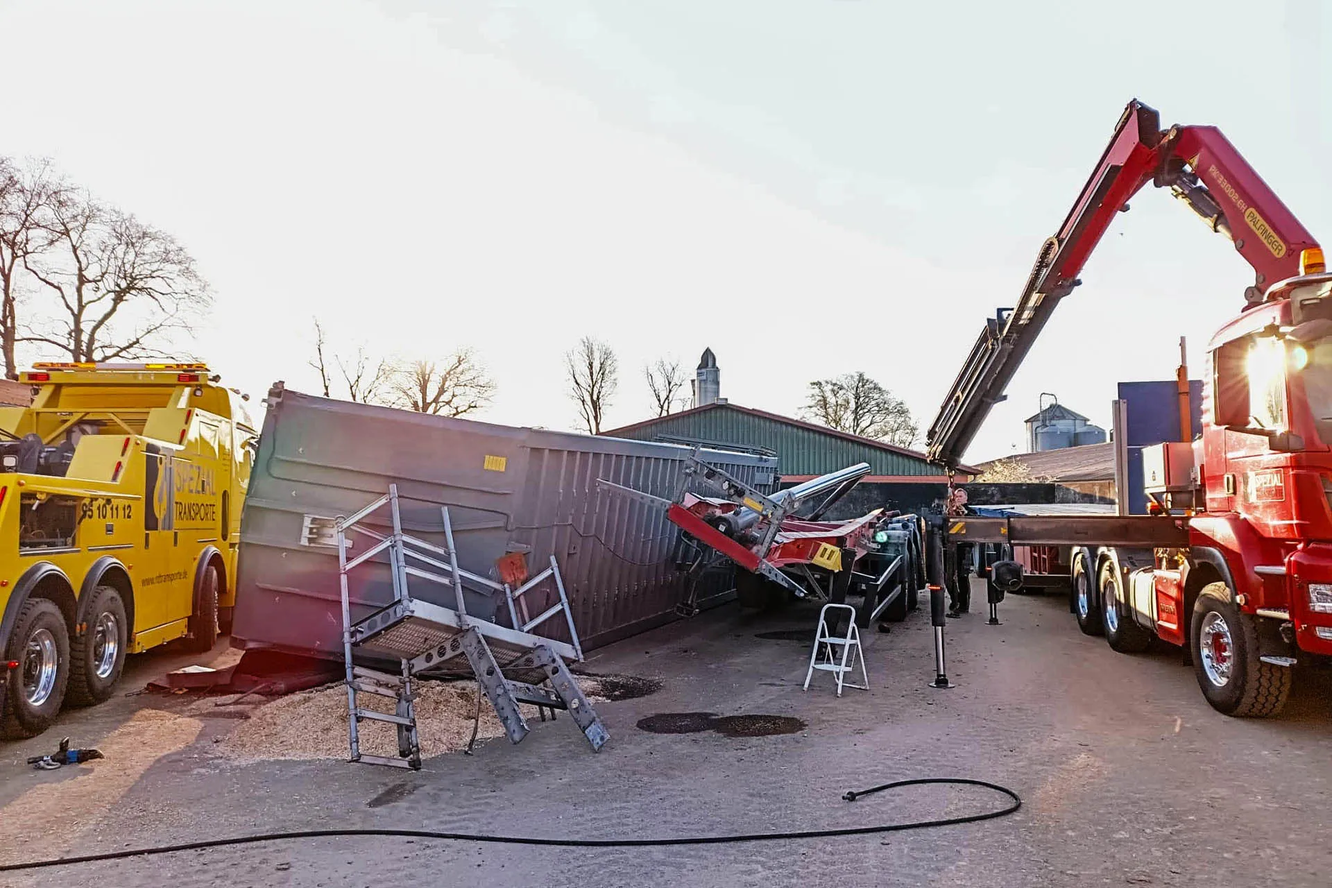 Bergungseinsatz von R.D. Spezial-Transporte: Ein gelber Abschleppwagen und ein roter LKW mit Ladekran sichern einen großen umgekippten Container auf einem landwirtschaftlichen Hof.