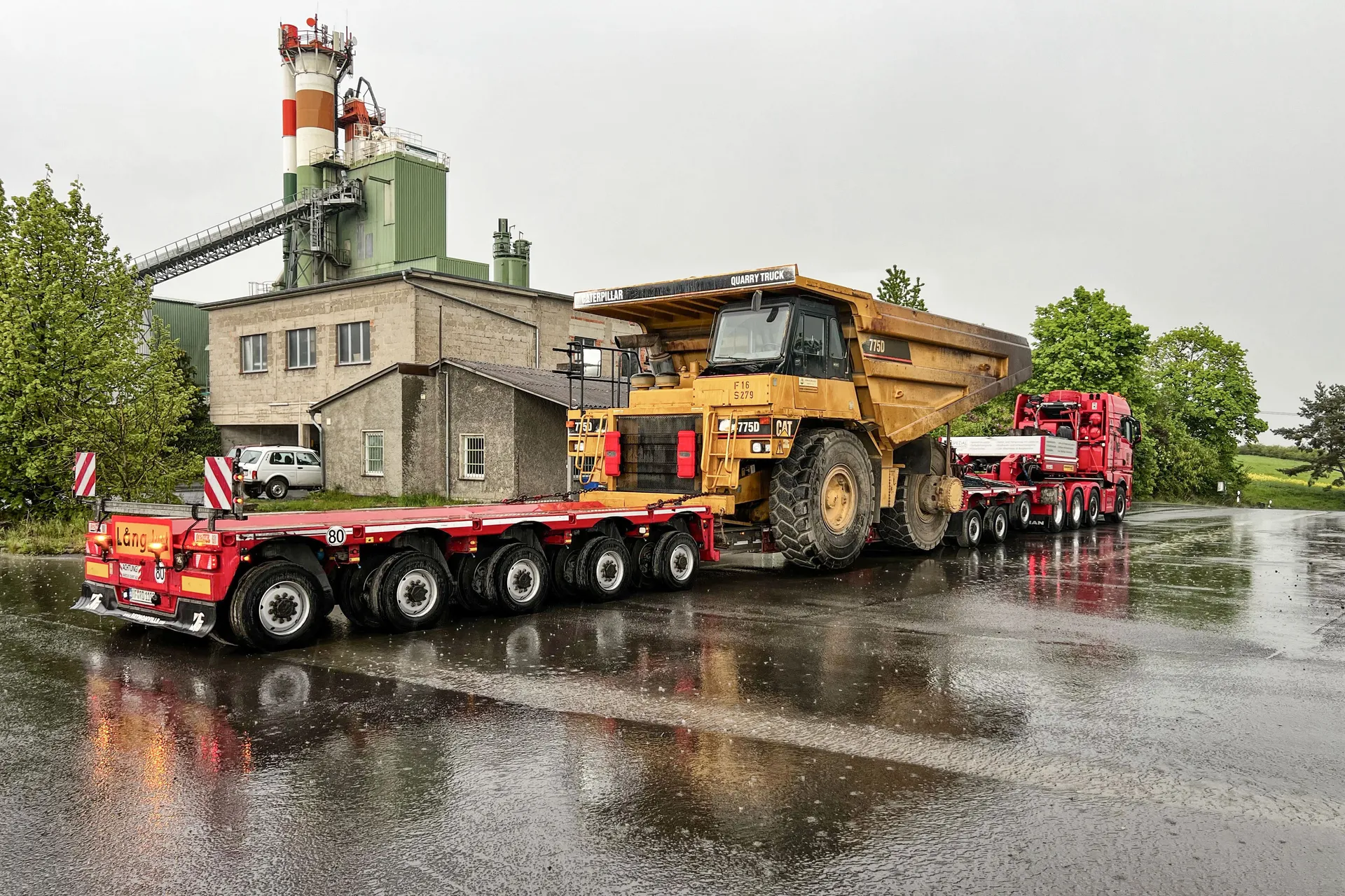 Schwertransport von R.D. Spezial-Transporte mit einem riesigen gelben CAT 775D Muldenkipper auf einem roten Tieflader, abgestellt auf nasser Fahrbahn vor einer Industrieanlage.