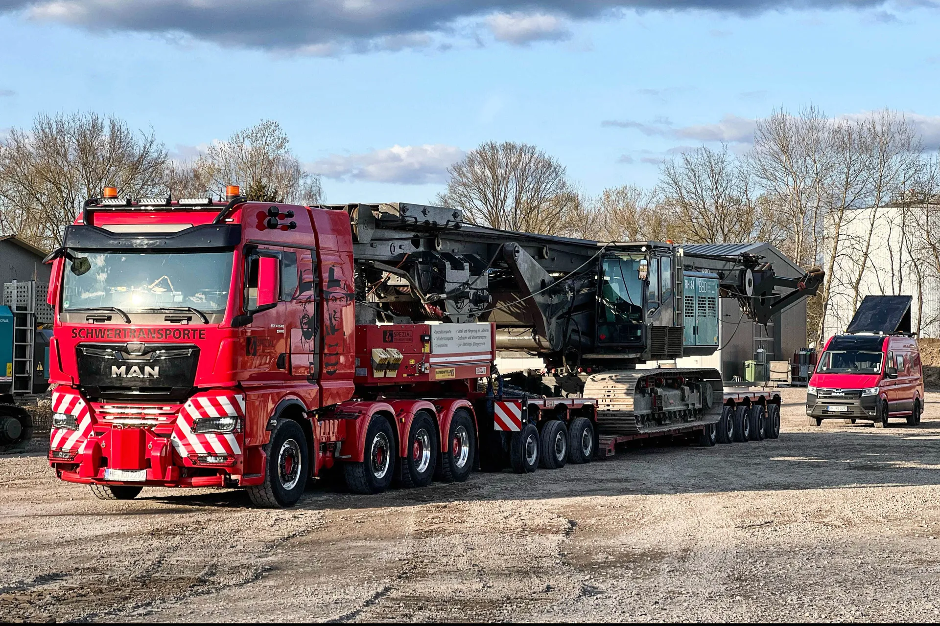Roter MAN Schwertransporter von R.D. Spezial-Transporte mit einer großen, schweren Industriemaschine auf einem Tieflader, begleitet von einem roten Service-Van auf einem Schotterplatz.
