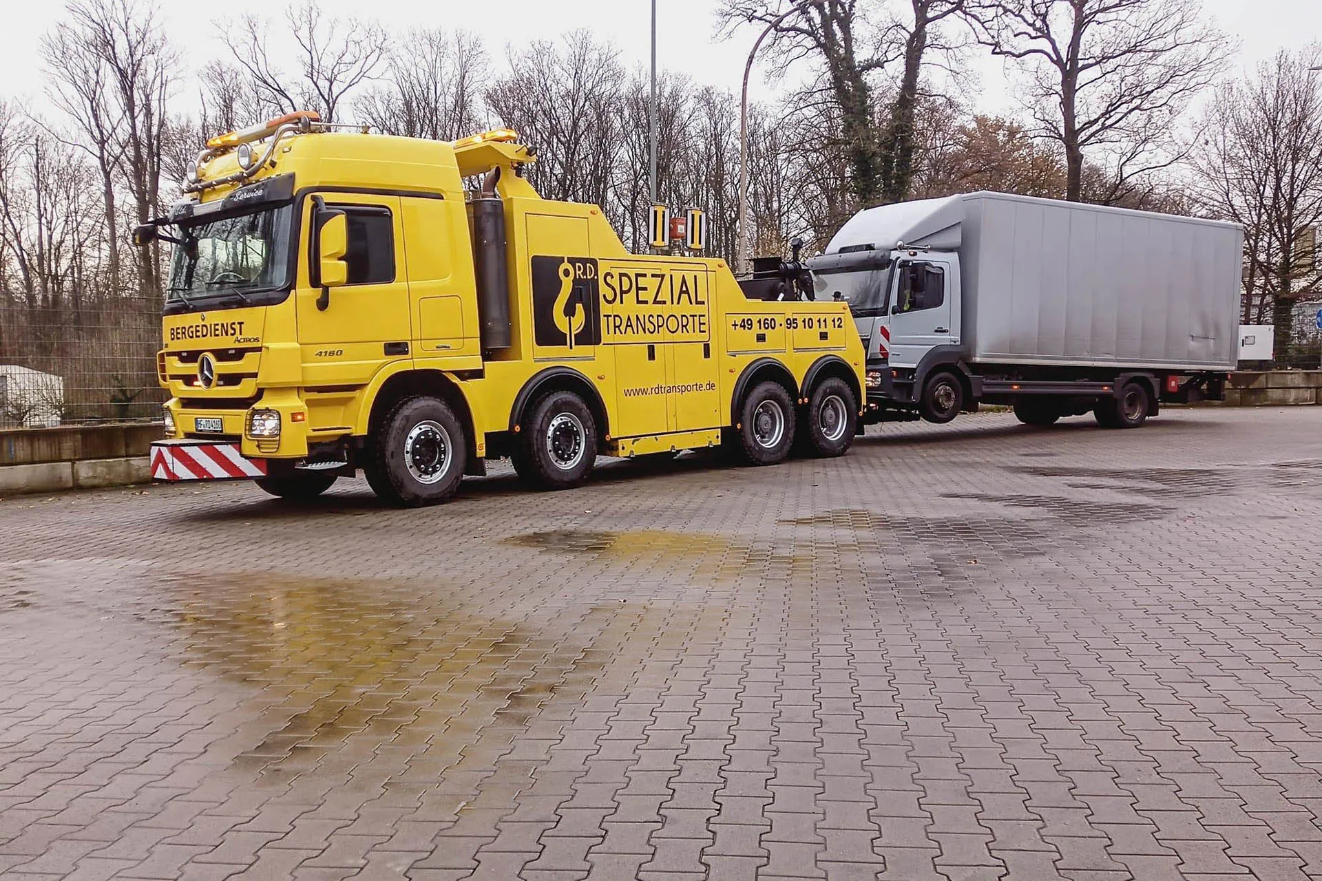 Gelber Mercedes-Benz Actros Bergedienst-LKW von R.D. Spezial-Transporte schleppt einen kleineren weißen Kastenwagen-LKW auf einem nassen, gepflasterten Hof ab.