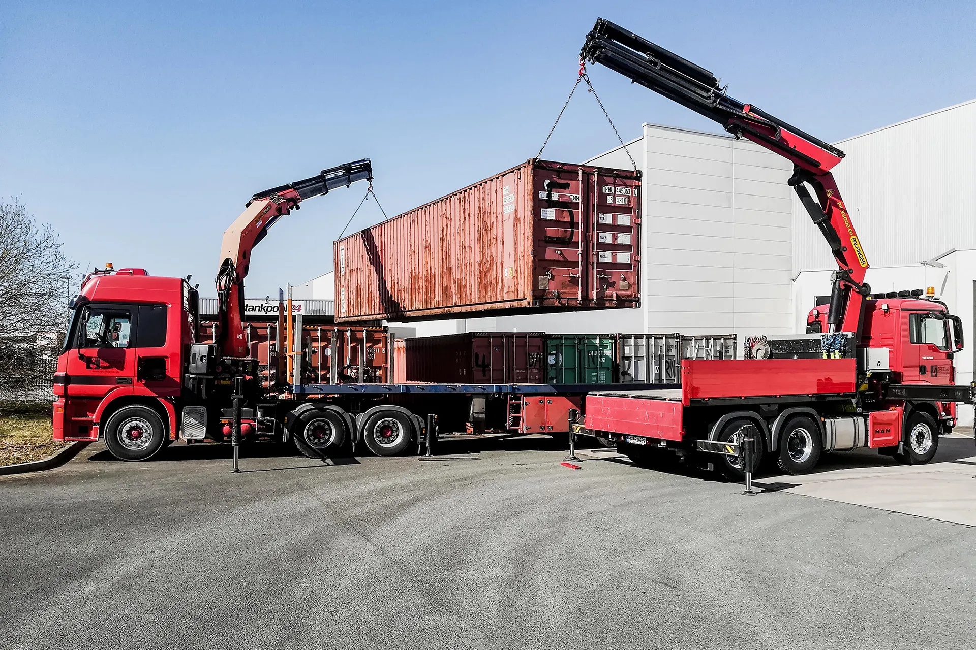 Tandemhub eines roten Schiffscontainers durch zwei rote LKW-Ladekrane von R.D. Spezial-Transporte auf einem Betriebsgelände.