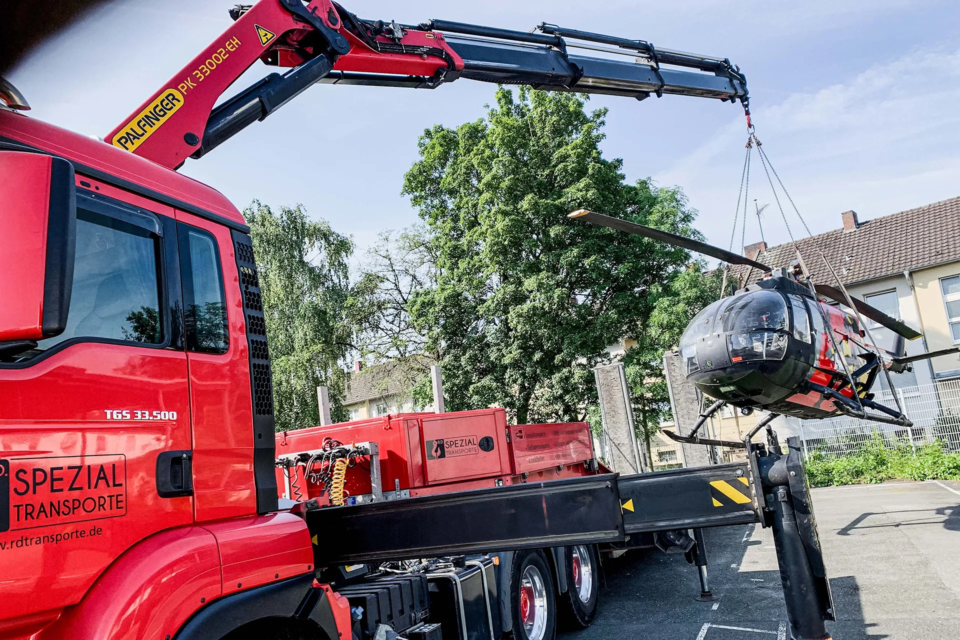 Seitliche Ansicht eines roten Spezial-LKW von R.D. Spezial-Transporte, der mit seinem Ladekran einen Helikopter anhebt.
