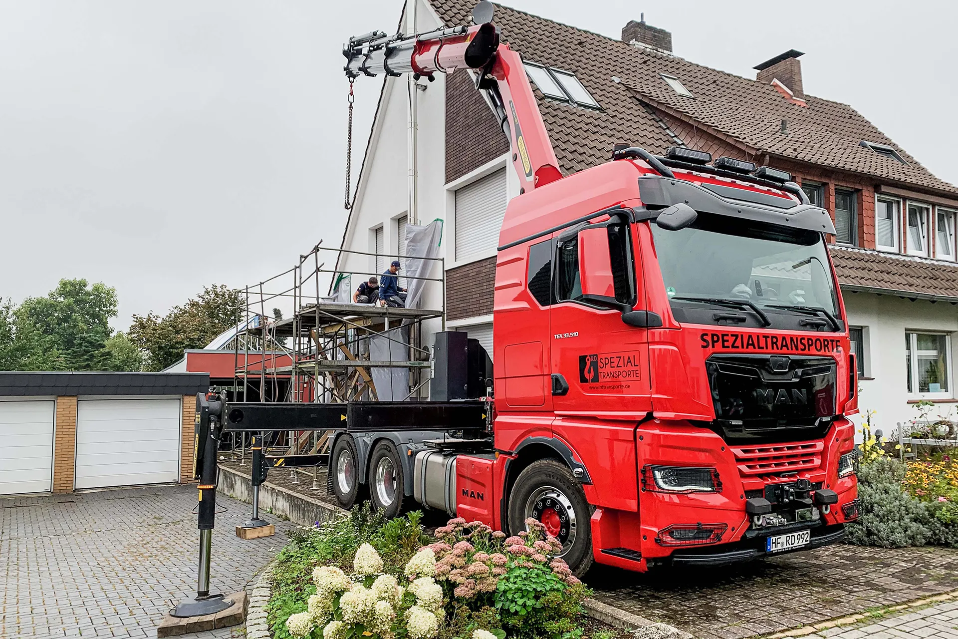 Roter MAN Spezial-LKW mit Ladekran im Einsatz vor einem Wohnhaus mit Baugerüst, vermutlich zur Materialanlieferung für Dacharbeiten.