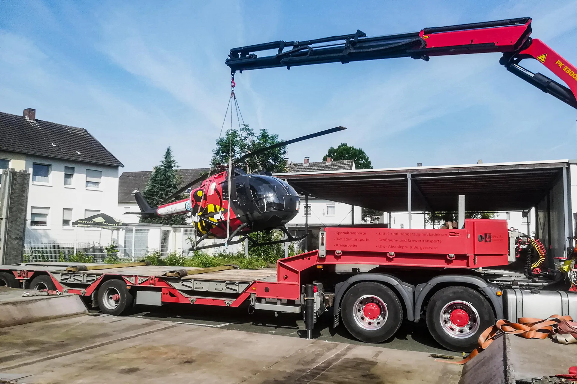 Verladung eines kleinen, rot-schwarzen Helikopters auf die Ladefläche eines roten Spezial-LKW mittels Ladekran.