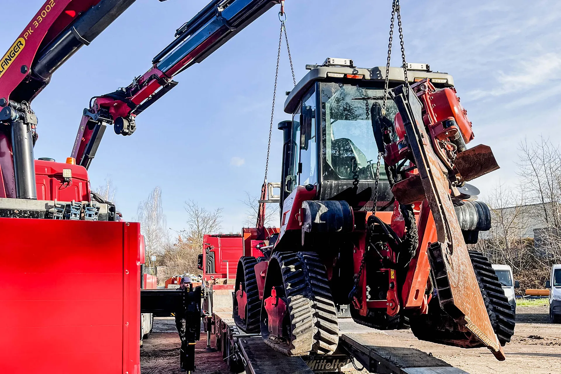 Verladung einer schweren, roten Forstmaschine oder eines Harvesters mit Greifarm auf einen Tieflader mittels Ladekran.