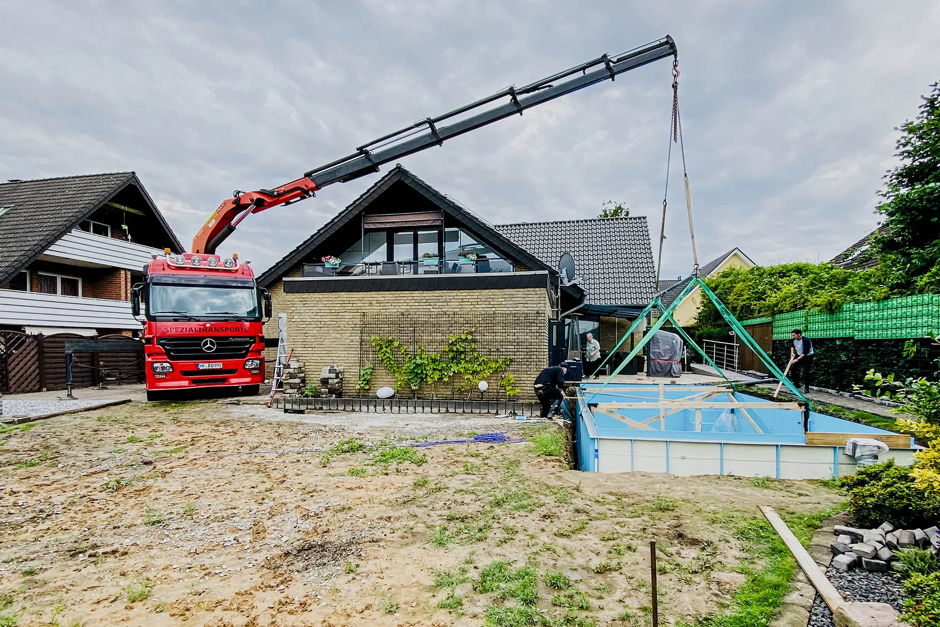 Ein roter LKW mit Ladekran hebt ein blaues Schwimmbecken-Element präzise in eine Baugrube in einem Hausgarten.
