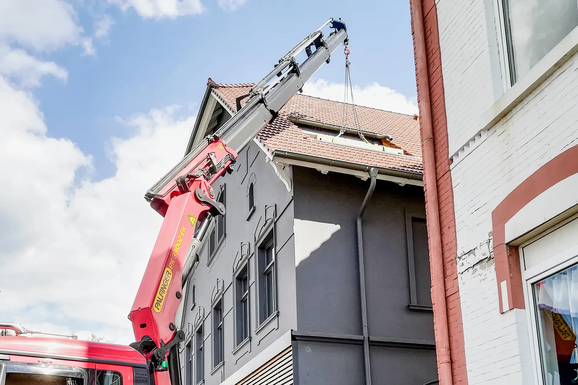 Ladekran eines roten Spezial-LKW hebt eine Last über das Dach eines Wohnhauses mit grauer Fassade und Ziegeldach.