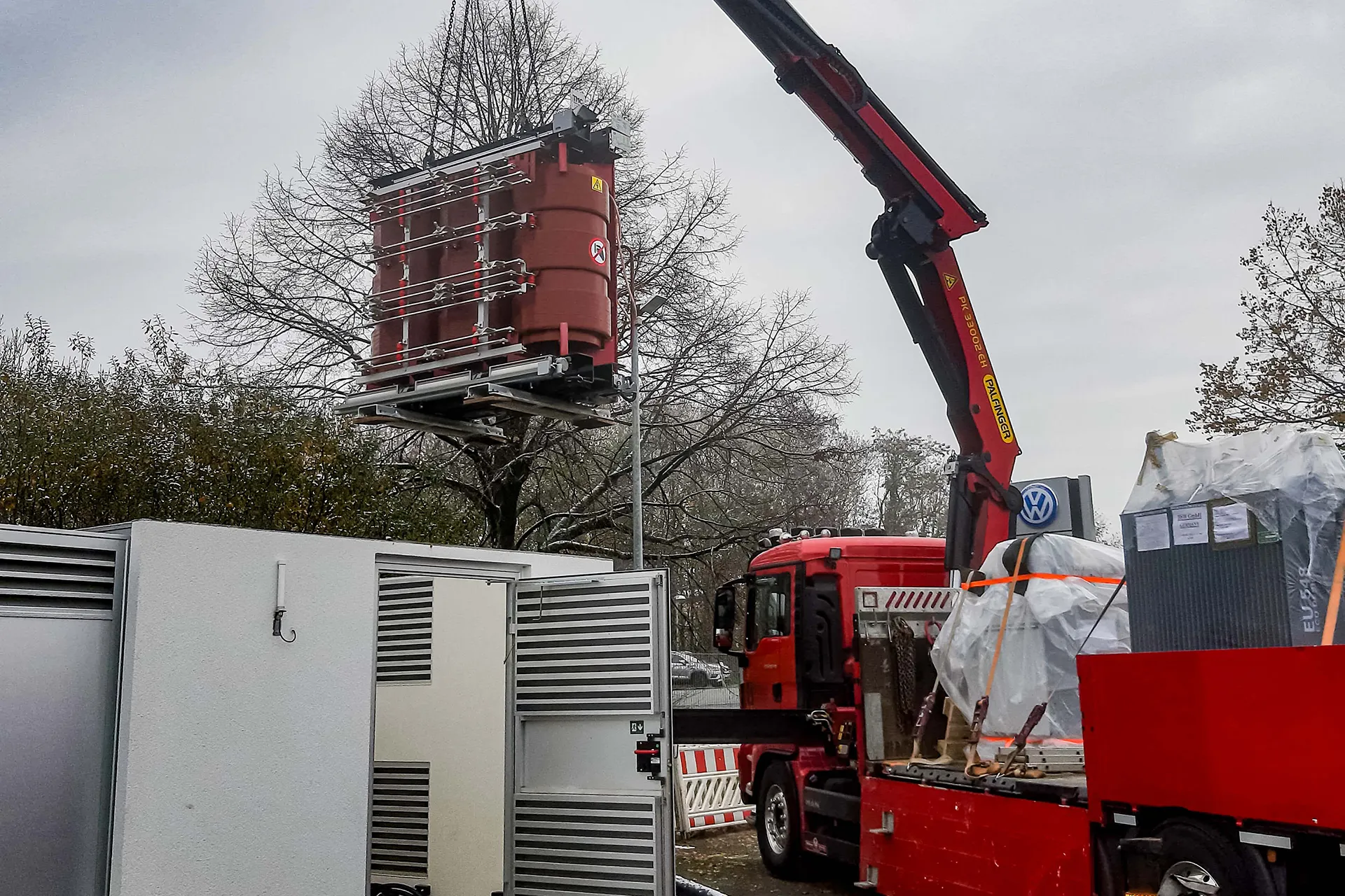 Ladekran hebt ein großes, braunes technisches Gerät, ähnlich einem Transformator, neben einem weißen Schaltkasten im winterlichen Umfeld.