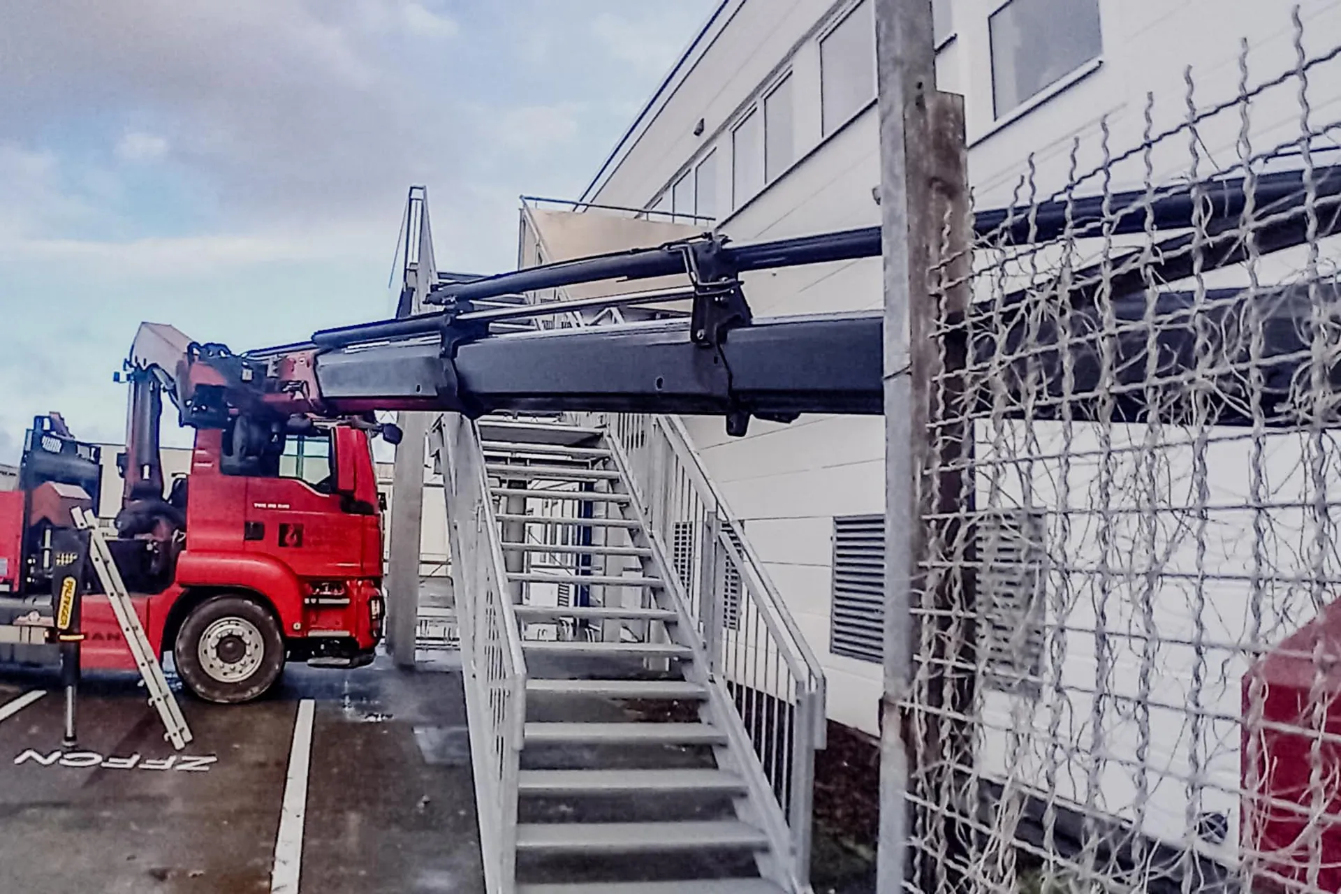 Roter LKW mit Ladekran manövriert präzise über eine metallene Außentreppe eines Gebäudes, um Material anzuliefern oder abzuholen.