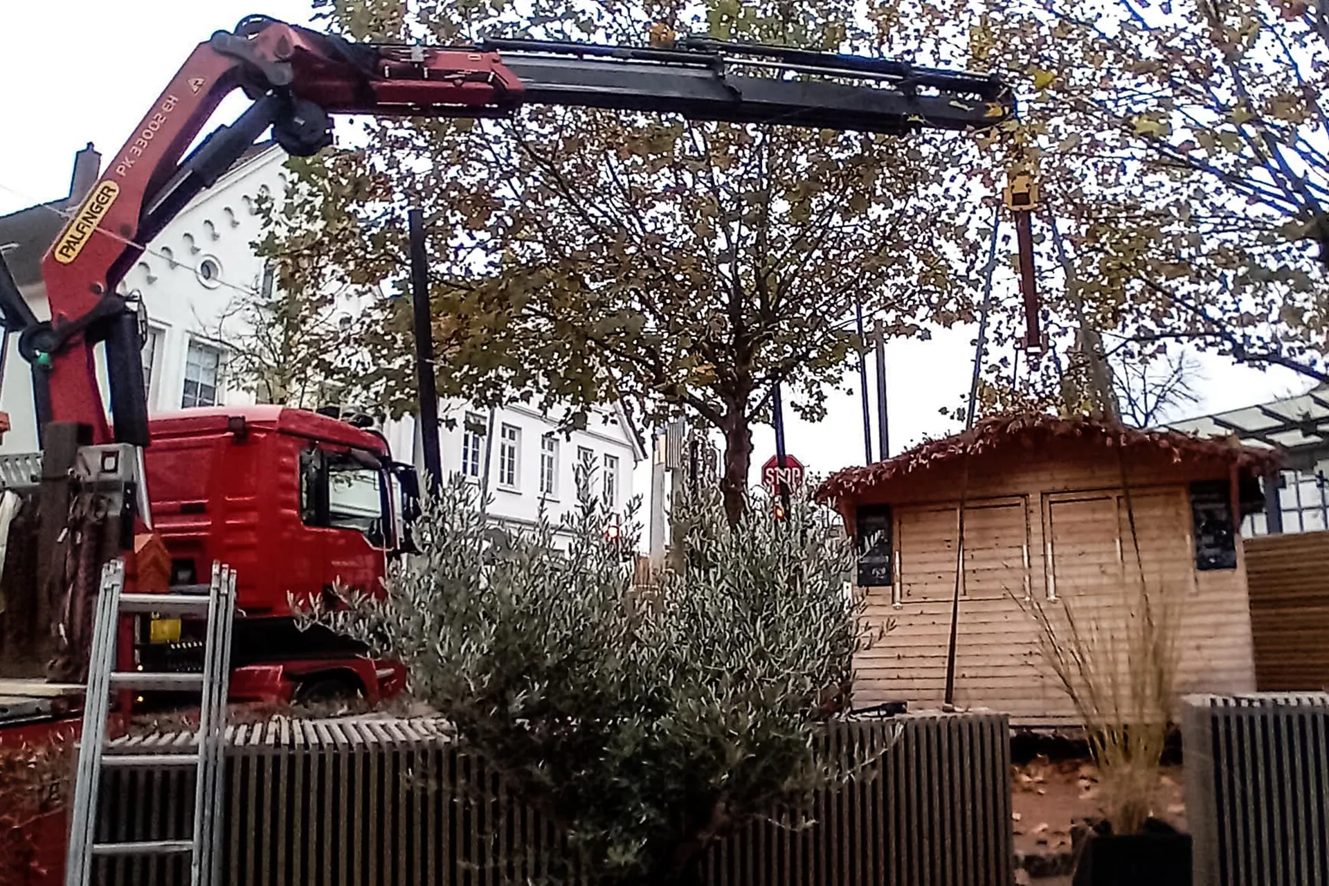 Ein roter LKW mit Ladekran positioniert eine Holzhütte oder einen Verkaufsstand auf einem städtischen Platz, möglicherweise für einen Markt oder eine Veranstaltung.