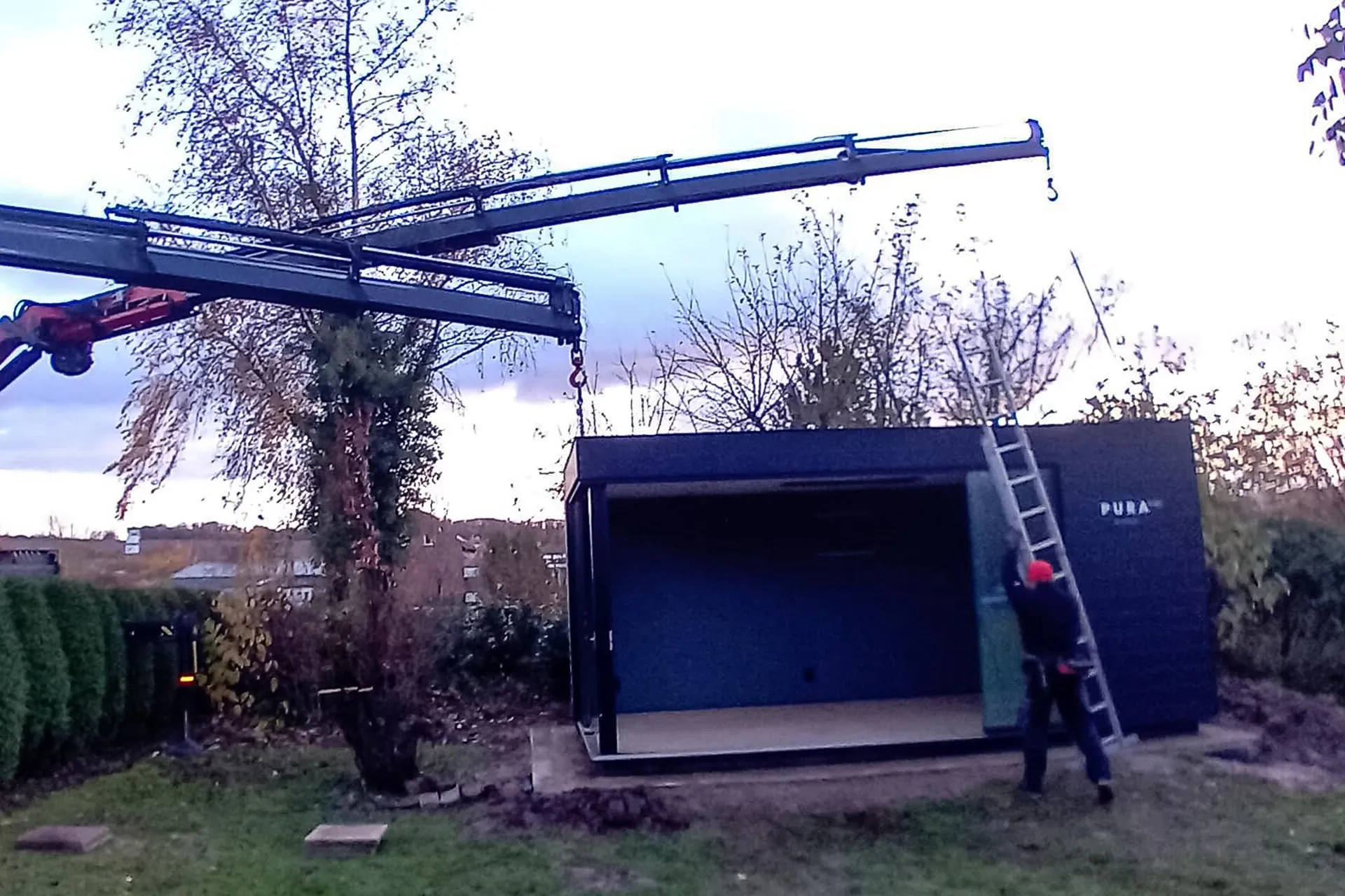 Ein Ladekran platziert ein dunkles, modernes Gartenhaus oder Modulgebäude in einem Garten, ein Arbeiter mit Leiter steht bereit.