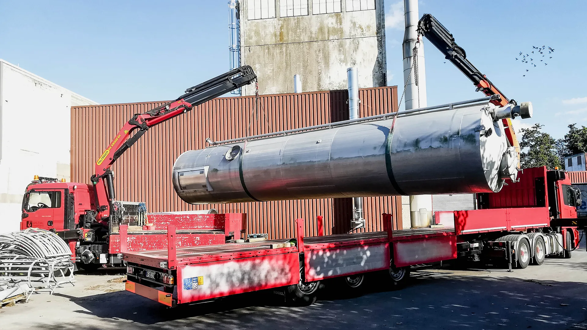 Präzise Kranarbeit: Zwei rote LKW mit Ladekranen positionieren einen langen, silbernen Industrietank auf einer Ladefläche vor einer Industriehalle.